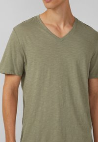 oliwkowa, krótka koszulka z dekoltem w serek, wykonana z miękkiego, teksturowanego materiału, z delikatnymi paskami i luźnym krojem.