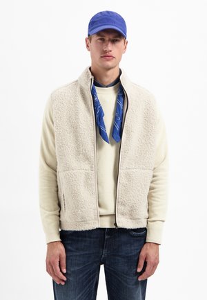 Mann mit blauer Kappe, beiger Fleeceweste über einem cremfarbenen Pullover, blauer Bandana um den Hals und dunklen Jeans, steht vor einem weißen Hintergrund.