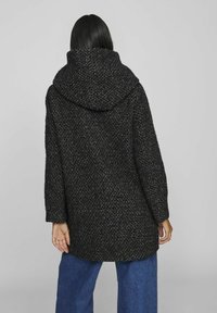 Manteau noir texturé avec une grande capuche et une coupe ample. Le tissu présente un motif tissé, rehaussé par des manches larges et une coupe droite.