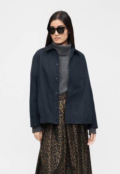 Žena s dlhými tmavými vlasmi, ktorá má na sebe čierne slnečné okuliare, tmavomodrú bundu cez sivý rolák a sukňu s leopardím vzorom na bielom pozadí.