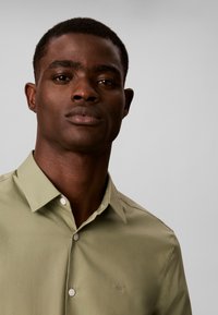 Homme aux cheveux courts portant une chemise boutonnée vert olive clair sur un fond gris uni.