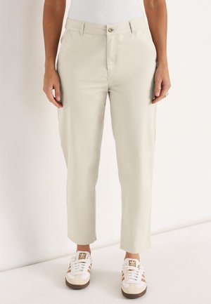 Lichtbeige rechte broek met knoop- en ritssluiting, gedragen met witte en beige Adidas sneakers en een witte top.