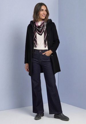 Une femme porte un manteau noir, une écharpe à motifs, un haut blanc, un jean foncé à jambes larges et des chaussures noires, sur un fond de studio bleu pâle.
