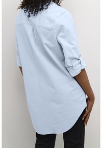 Chemise bleu clair à manches longues avec poignets retroussés, pli dans le dos, ourlet arrondi et texture douce. Convient pour une tenue décontractée ou semi-formelle.
