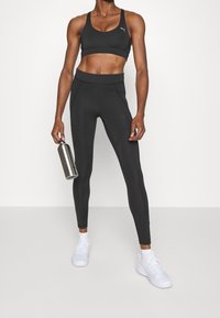 Svart sport-BH och högmidjade leggings i en åtsittande design, ihop med vita sneakers. En metallisk vattenflaska hålls i höger hand.