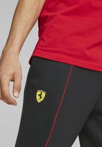 Černé sportovní kalhoty s červeným bočním pruhem, zdobené žlutým logem Ferrari. Tričko je červené s hladkým textilním povrchem.