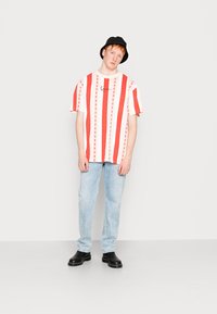 Rød og hvid stribet t-shirt med korte ærmer, der har et centralt logo, parret med lyseblå jeans og sorte sko. Sort bøttehat.