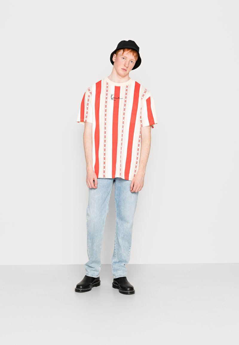 Rød og hvid stribet t-shirt med korte ærmer, der har et centralt logo, parret med lyseblå jeans og sorte sko. Sort bøttehat.