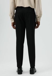 Zwarte, op maat gemaakte broek met een smalle pasvorm, voorzien van twee achterzakken en een gladde stof, gecombineerd met een beige shirt met lange mouwen.