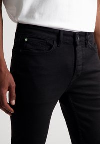 Jeans de mezclilla negra con corte slim, que cuentan con bolsillos frontales, detalles de costura sutiles y un pequeño acento verde en la cintura.