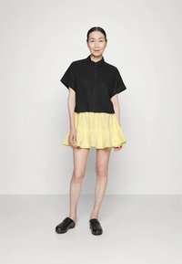 Camisa preta de botão de manga curta combinada com uma saia mini amarela com babados e tamancos pretos. Tecido suave com cores contrastantes.