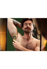 Un uomo con baffi e orecchino a cerchio sta usando un rasoio giallo e nero sulla sua ascella. Ha un tatuaggio e la sua pelle  è di tonalità chiara.