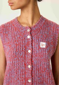 Detailný záber na osobu v bezrukávovom červenom a modrom pletenom kardigane s bielymi gombíkmi a nálepkou "LA MAISON AMV" na hrudi.