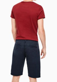 Muž v červené košili s krátkým rukávem a tmavě modrých kraťasech po kolena stojí se zády k fotoaparátu.