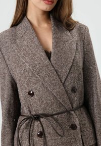 Bruine blazer met dubbele knoopsluiting en visgraatmotief, voorzien van een ceintuur, grote donkere knopen en een zachte textuur.