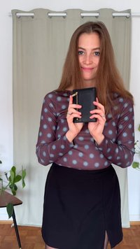 Black textured wallet with a gold zipper, held by a person in a polka dot long-sleeve top and a black skirt. Natural light background.