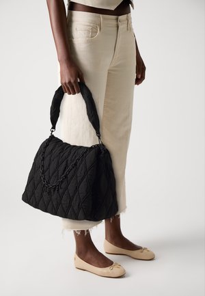 Person holding a large black quilted handbag with chain detail, wearing cream high-waisted jeans and beige ballet flats.