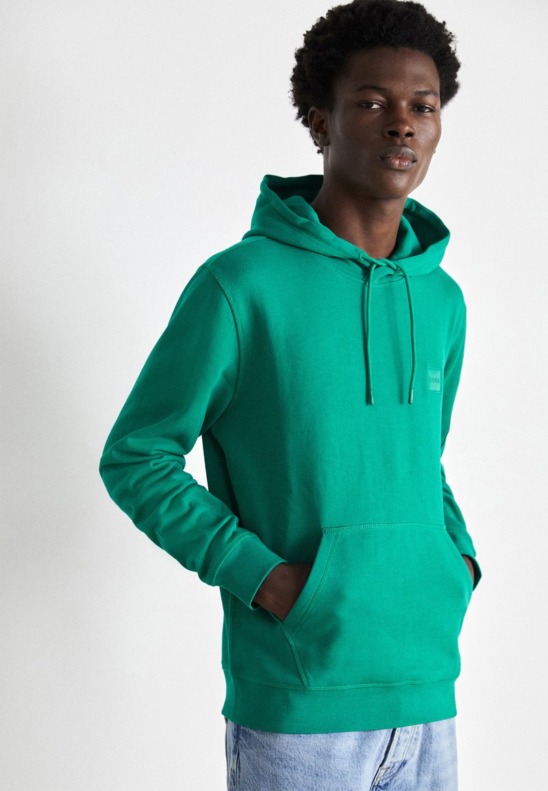 Green hoodie made of cotton blend fabric, featuring a drawstring hood, front pocket, and ribbed cuffs. Simple design with textured finish.