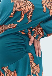 Abito in tessuto turchese con stampe di tigri arancioni, con dettagli arricciati sui lati e maniche morbide. Texture morbida e liscia.