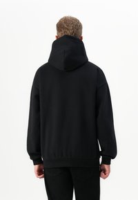 Homme aux cheveux courts et bouclés portant un sweat à capuche noir et un pantalon noir, debout face à un fond blanc, de dos.