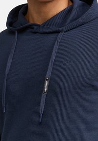 Sudadera de color azul marino confeccionada en tejido acanalado, con cierre de cordón y un pequeño logo en el pecho. Detalle de superficie texturizada visible.
