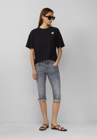 Schwarzes Baumwoll-T-Shirt mit kurzen Ärmeln und Sonnensymbol, kombiniert mit hellgrauen hochgekrempelten Jeans und schwarzen flachen Sandalen.