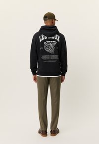 Μαύρο φούτερ με κουκούλα με λευκό γραφικό και κείμενο στην πλάτη, συνδυασμένο με ελαιδί παντελόνι και καφέ παπούτσια. Το μοντέλο φαίνεται από πίσω.