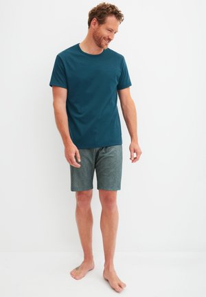 Mann trägt ein türkisfarbenes Kurzarmhemd und gemusterte, grüne Shorts bis zum Knie, steht barfuß vor weißem Hintergrund und schaut lächelnd nach unten.