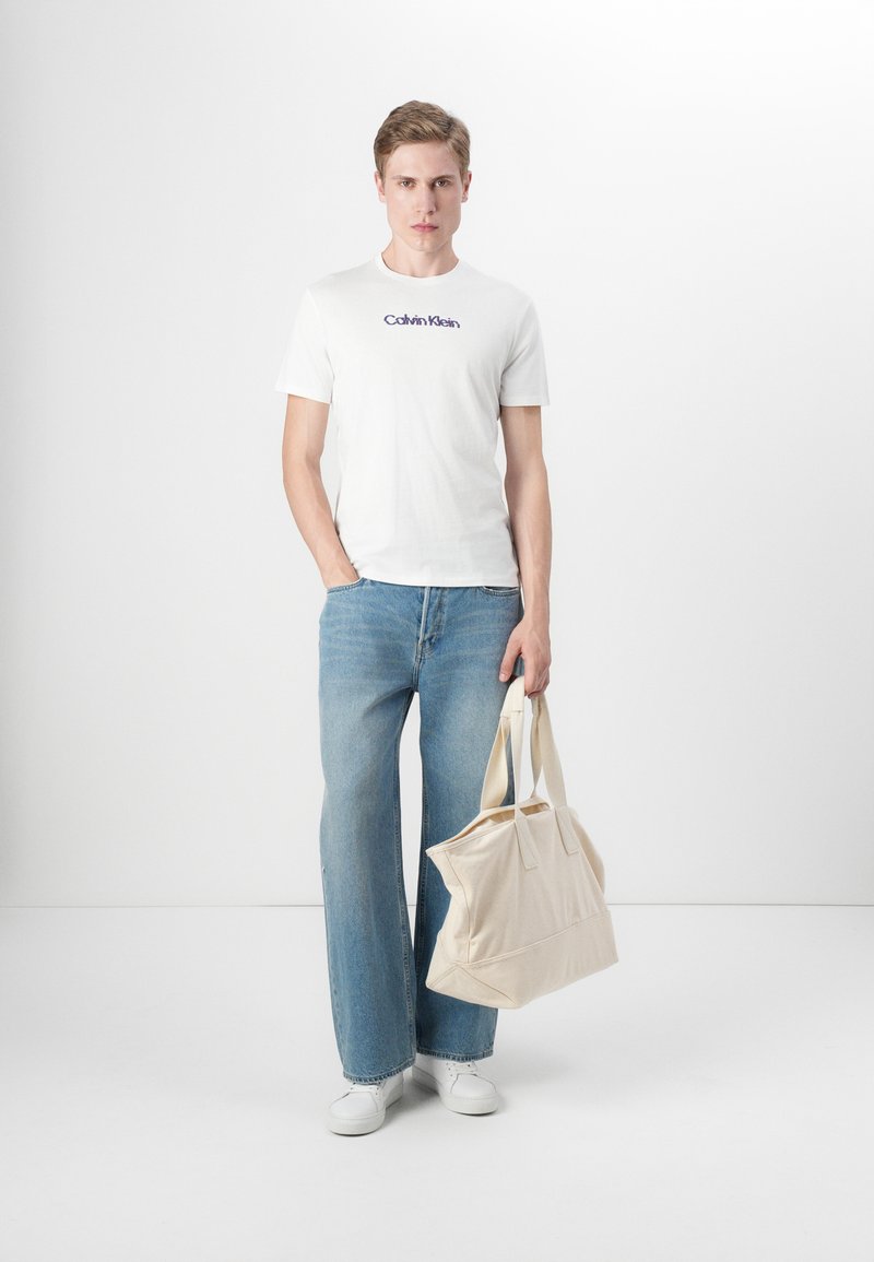 Camiseta blanca de algodón con el logo azul de "Calvin Klein", jeans de pierna ancha azules, bolso tote beige y zapatillas blancas en un entorno minimalista.