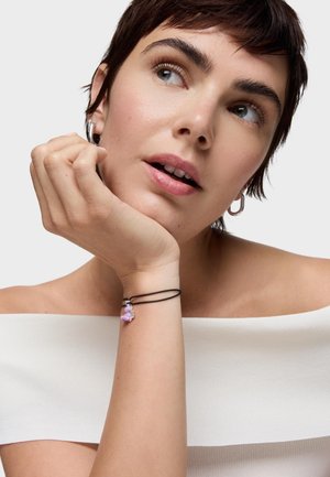 Młoda osoba z krótkimi ciemnymi włosami, ubrana w biały top odsłaniający ramiona, srebrne kolczyki koła, opierająca podbródek na ręce z czarną bransoletką i różowym zawieszonym charms.