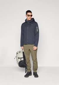 Navy jacket with a hood and zippered pocket, paired with light green pants and black hiking shoes. A gray and black backpack is held in one hand.