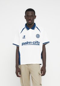 Λευκό πόλο πουκάμισο με σκούρες μπλε λεπτομέρειες και γιακά, με κείμενο και λογότυπο "palm city" στο μπροστινό μέρος, κατασκευασμένο από υφασμάτινο υλικό με υφή.