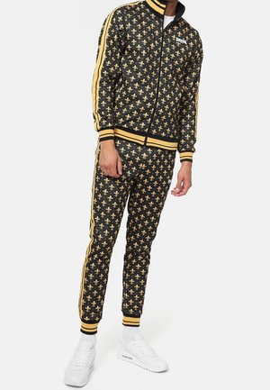 Homme portant un survêtement noir et or à motifs avec imprimé fleur-de-lis et bandes jaunes, associé à des baskets blanches.