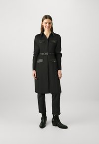 Zwarte shirtjurk met lange mouwen, zakken en een ceintuur in de taille. Gecombineerd met donkere jeans en zwarte enkellaarsjes. Glad stoftextuur.