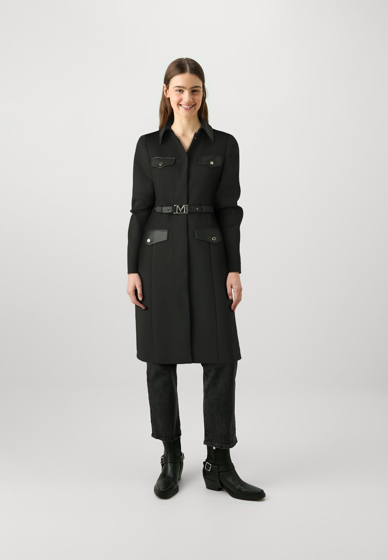 Zwarte shirtjurk met lange mouwen, zakken en een ceintuur in de taille. Gecombineerd met donkere jeans en zwarte enkellaarsjes. Glad stoftextuur.