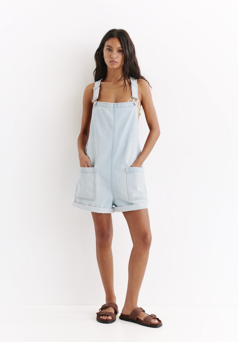 Jeune femme debout contre un mur blanc, portant une salopette courte en jean bleu clair et des sandales marron à boucle, mains dans les poches, expression neutre.