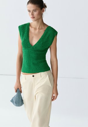 Femme portant un haut sans manches texturé vert à col en V profond et un pantalon large crème, tenant une petite pochette grise tissée.