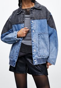 Chaqueta vaquera con diseño a bloques de color en paneles gris oscuro y azul claro, cierre frontal con botones y dobladillo acanalado texturizado.