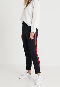 Pantalones de chándal azul marino con una franja roja lateral, cinturilla elástica y puños, combinados con una camiseta de manga larga blanca y zapatillas blancas.
