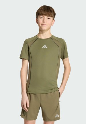 Tinejdžer stoji s jednom rukom u džepu, nosi maslinasto zelenu sportsku majicu i odgovarajuće šorceve s bijelim Adidas logotipima.