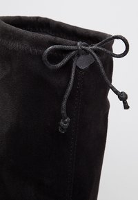 Bottine en daim noir présentant un dessus arrondi, un laçage décoratif et une texture lisse. Le laçage est tressé et noué en un nœud.
