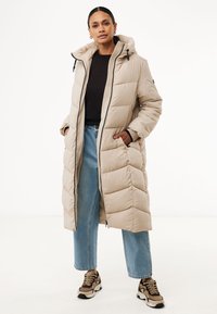 Lange beige pufferjas met een capuchon, voorzien van een verticale gestikte ontwerp, ritssluiting en zijvakken. Combinatie met blauwe jeans en sneakers.