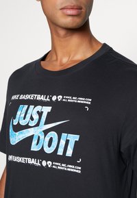 Camiseta de algodón negra con cuello redondo, que presenta un gráfico en azul y blanco con el lema "JUST DO IT" junto con la marca Nike y detalles de baloncesto.