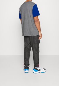 Camiseta de color bloque en gris y azul, pantalones cargo negros con bolsillos y zapatillas negras y blancas con detalles en azul. Conjunto casual visto desde atrás.