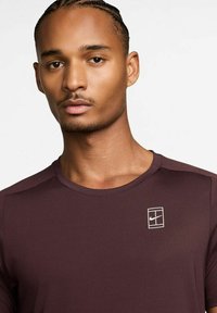 Burgundska športna majica z okroglo ovratnikom, gladko teksturo in belim logotipom Nike na levem prsnem delu. Minimalističen in pristen dizajn.