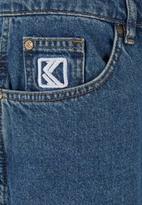 Mėlyni džinsai su užpakaline kišene, kurioje yra išsiuvinėtas baltas logotipas. Siuvinėjimas yra matomas, su vario atspalvio detalėmis.