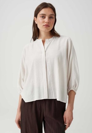 Femme portant un chemisier blanc texturé ample avec des manches élastiques trois-quarts et un pantalon large sombre, debout devant un fond clair uni.