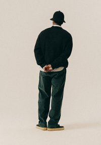 Μαύρο πλεκτό πουλόβερ, χαλαρές πράσινες παντελόνες και μαύρο καπέλο τύπου bucket. Το μοντέλο στέκεται με τα χέρια σφιγμένα πίσω από την πλάτη, στραμμένο μακριά από τον θεατή.