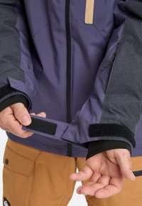 Person adjusting the velcro cuff of a purple and gray jacket, wearing tan pants, with a black inner wrist layer visible.