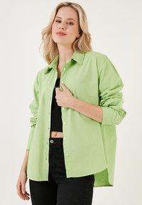 Camicia verde chiaro a maniche lunghe con chiusura a bottoni, colletto alla francese e maniche arrotolate, abbinata a una maglietta nera e pantaloni neri.
